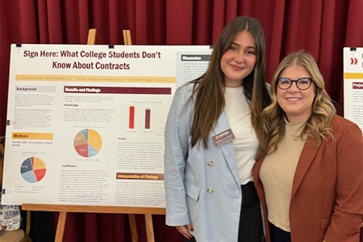 Carmella Cortez stands with her advisor, Jill Lauderman, in front of a poster displaying Carmella’s honors research. Carmella has long brown hair and wears a white shirt and light blue blazer; Jill has long blond hair and wears a burnt orange blazer.