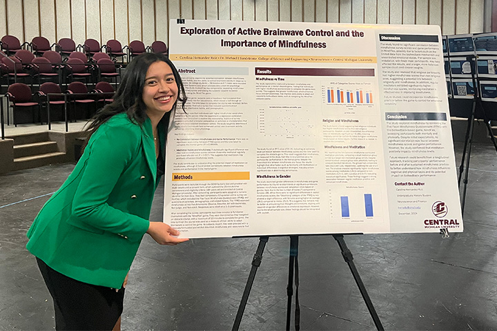 Carolina Hernandez Ruiz wear a green blazer and black skirt and poses with her honors research poster on brainwave control and mindfulness..