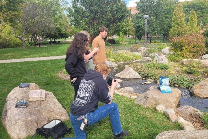Four students stand around a game box for Hydrologic Cycle which has been posed on a rock in the outdoor gardens on the CMU campus. The students have cameras and phones and are taking photos for the game's publisher.