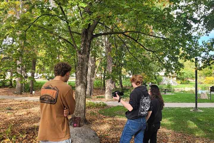 Three students stand outside on the campus of CMU. They are under a tree and have set the game Eyeball to Eyeball on a rock to photograph it for a class project. One student holds a camera.