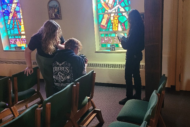 Three students gather in St. Mary's Catholic Parish. They has posed a the game box for Monumental Consequence in front of a stained glass window. On student sits in a chair looking at the viewfinder of their camera.