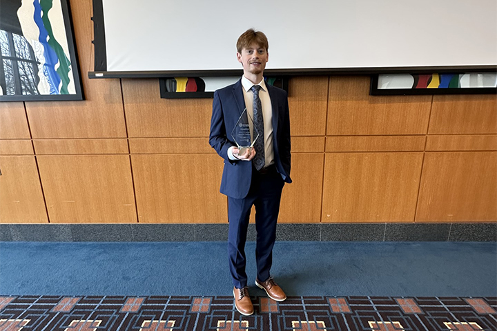 Cole Carpenter stands in a suit holding a crystal plaque after a finance competition, posing in front of a presentation screen.