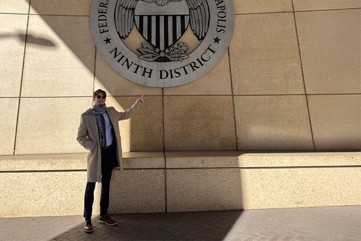 Cole Carpenter stands outside the Federal Reserve Bank of Minneapolis, wearing a coat and scarf, pointing toward the Ninth District seal on the building wall.