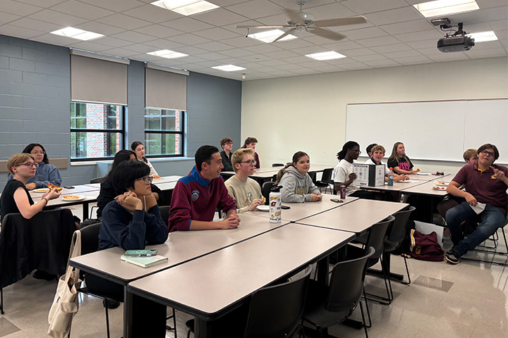 Students sit at classroom tables with pizza and drinks, looking toward someone outside the frame.