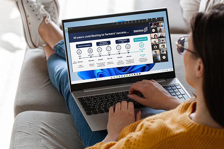 A student sits on a couch with a laptop on her lap. On the screen is a PPT slide with content from Cristiano Machado Costa's presentation. Students appear in the participant gallery on the screen as well.