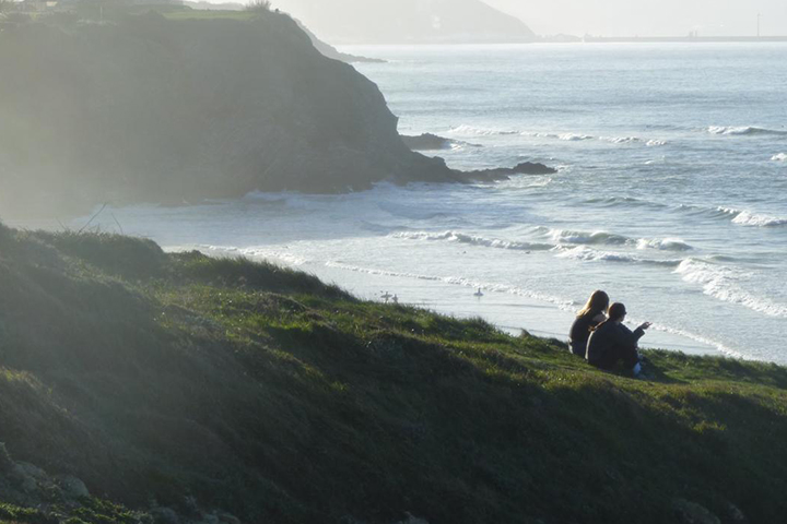 Ella See sits with a friend on a seaside cliff in Bilbao spain. The photo is taken from a distance, so they sit in a green grassy field that overlooks the water below.