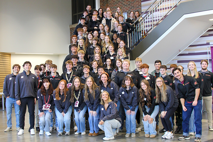A large group of high school students from Sacred Heart and CBA mentors crowd the staircase in Grawn Atrium. They all wear black long-sleeve CMU Next-Gen logo gear. Some students wear large plastic maroon and gold chains with Action C pendants.