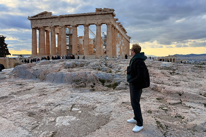 CMU student Ethan Rauschert stands on the rocky Acropolis in Athens, Greece, with the ancient Parthenon behind him.