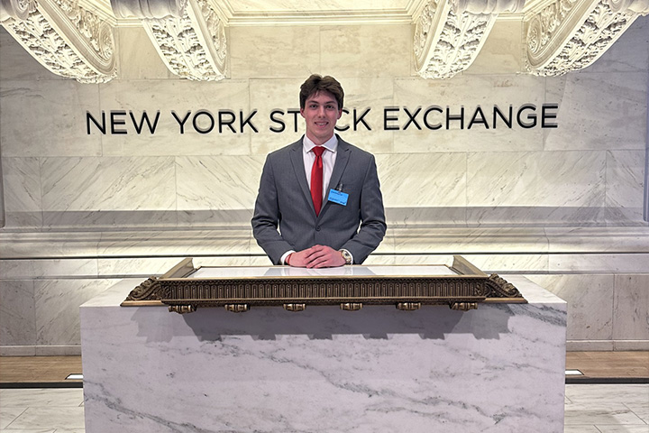 Ethan Schweitzer, a finance and logistics management student, stands at the New York Stock Exchange in a gray suit and red tie.
