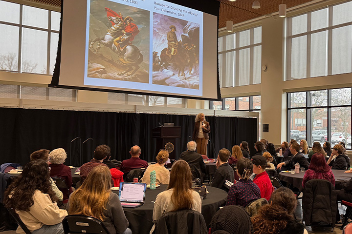 Joanne Ciulla stands behind a podium on a raised stage in the Grawn Atrium during the 2026 Ethics Summit. Behind her is a screen with portraits of Napoleon. In the foreground, students sit at round tables and listen intently.