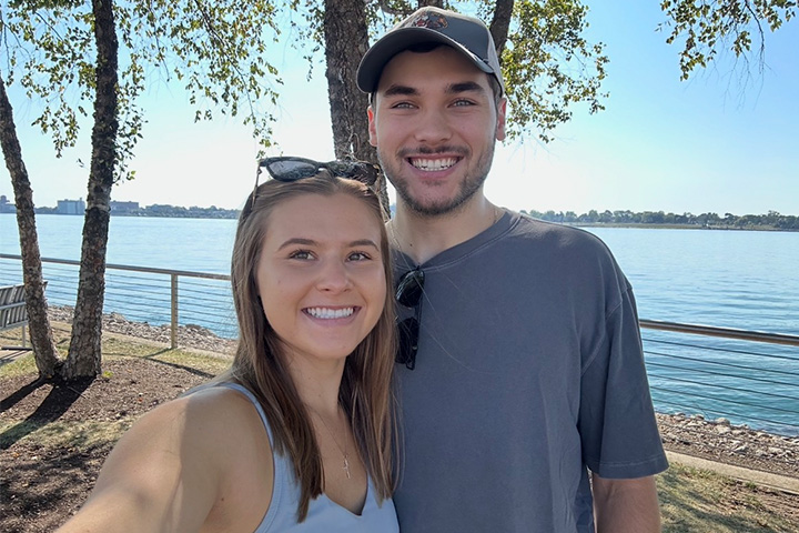 CMU graduates Francis Serazio and Abby Kuczmarski stand together at the edge of a lake. They have on summer clothes and each are carrying sunglasses.
