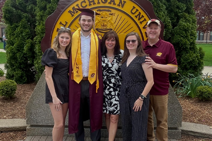 Francis Serazio stands in his cap and gown with his family in front of the CMU seal on Warriner Mall.