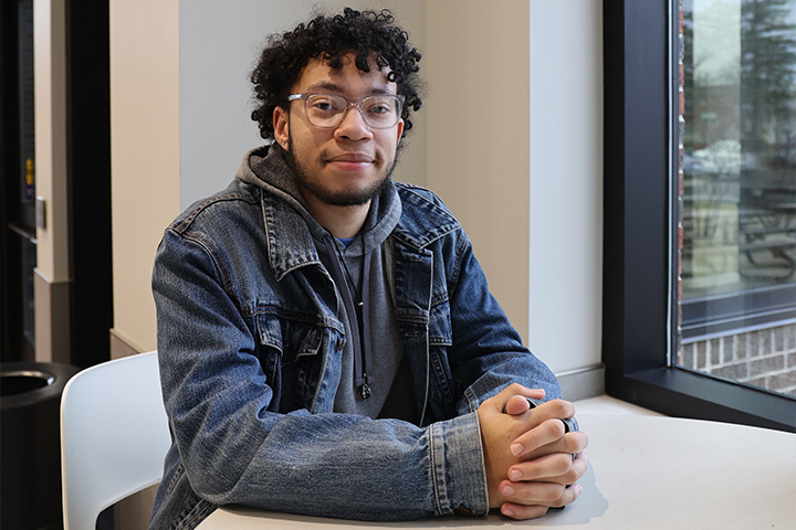 Garin Turner sits at a table inside a campus building, facing the camera with his hands clasped. Natural light from a large window illuminates the scene, with campus walkways and trees visible outside.