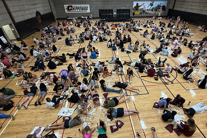 Students crowd a basketball court as they group up to playtest a new CMich Press board game. Many are laying in circles or sitting in groups looking at game components and working as teams to playtest the game.