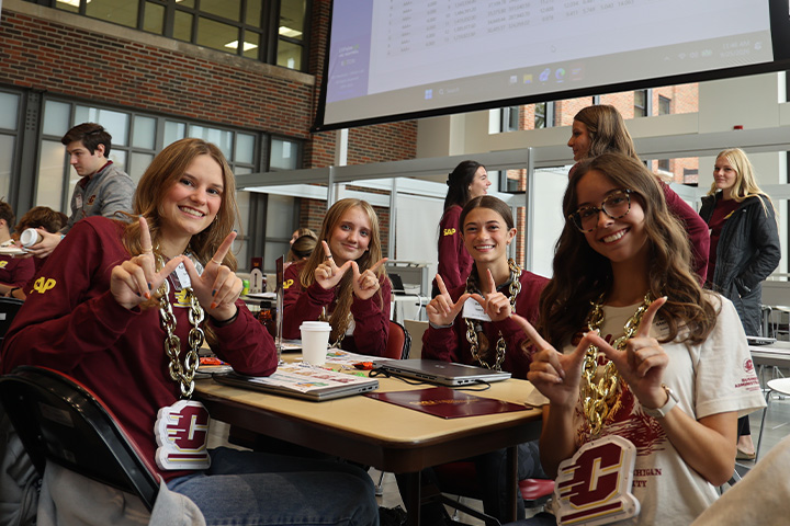 Four ERPsim participants smile and make CMU hand signs at a table with laptops, wearing gold chains with CMU emblems.