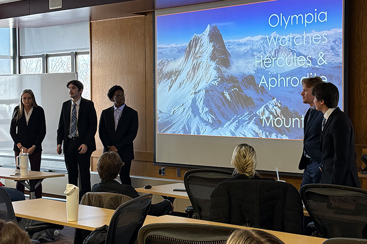 Five students stand at the front of a conference room wearing business professional clothing. They are presenting their iCore project. Behind them in a display screen with a PPT slide showing their smartwatch brand.