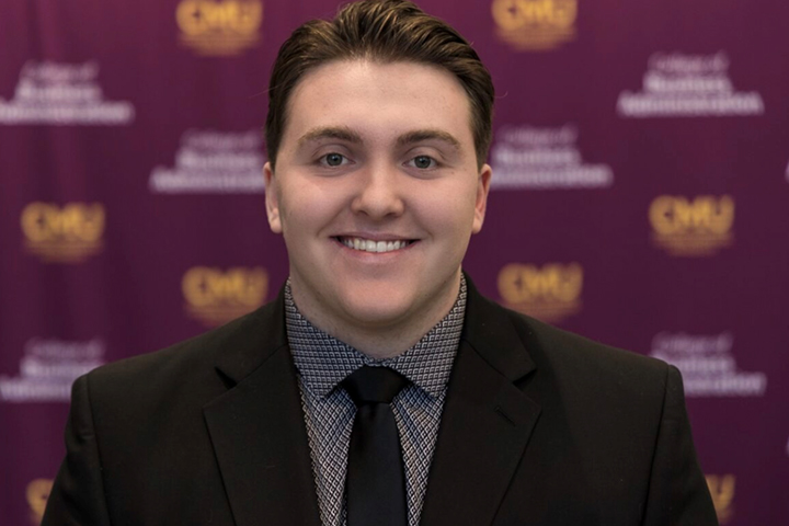 Jackson Raymond wears a black suit jacket, a dark gray black patterned shirt with a black tie. He has brown hair that is combed back.