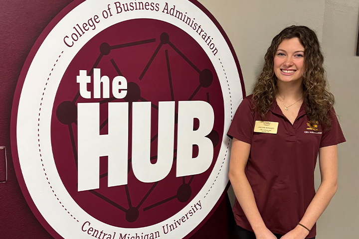 Jaclyn Serazio wears a CMU polo shirt with a name tag and stands by a HUB logo.