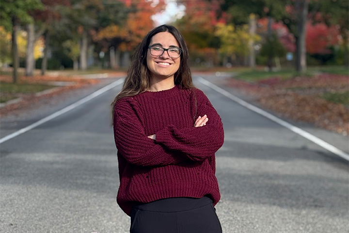 Jade Walleman stands confidently in the middle of a quiet road lined with colorful autumn trees, smiling with her arms crossed while wearing a burgundy sweater and black pants.