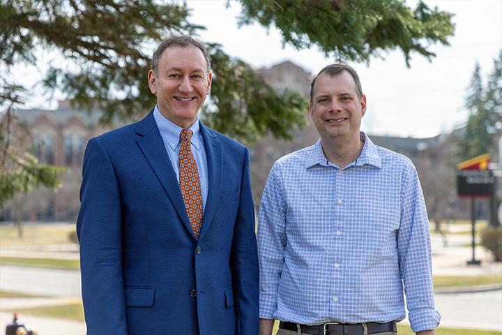 Adam Epstein (left) and Jason Taylor stand outdoors on Central Michigan University’s campus, smiling at the camera with campus buildings and trees in the background.