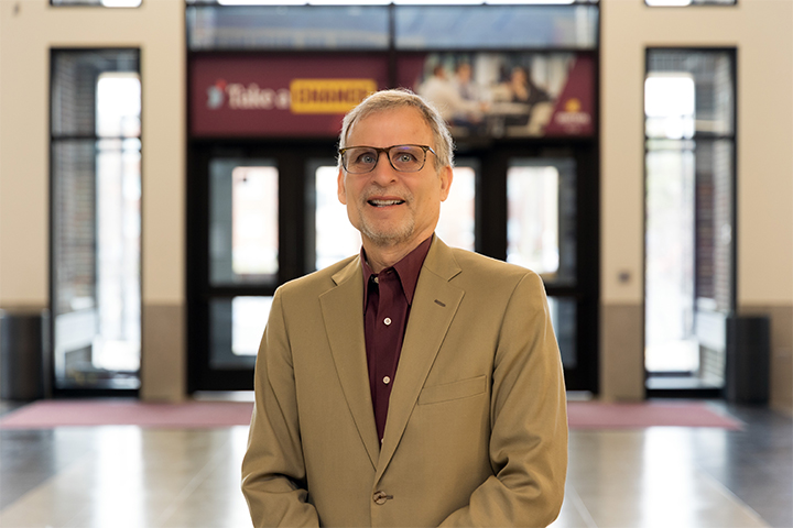Jeffrey Hoyle, marketing and professional sales faculty member at Central Michigan University, stands smiling in the Grawn Hall Atrium wearing a tan blazer and maroon shirt.