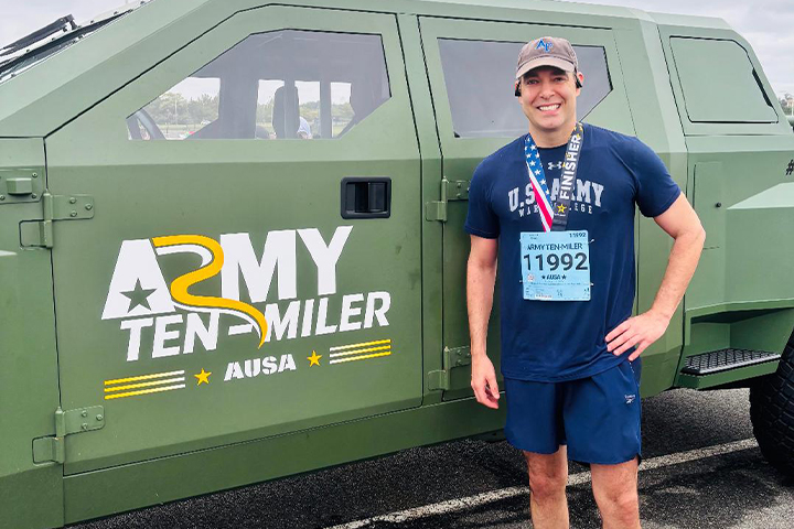 Jeremy Jackson wears his running clothes with a race bib and finisher medal for the Army Ten Miler. Behind him is a branded army vehicle with a logo for the race.