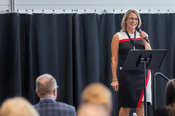 Julie Messing stands at a podium speaking into a microphone during a College of Business Administration event at Central Michigan University. Audience members are visible in the foreground as she addresses the crowd.