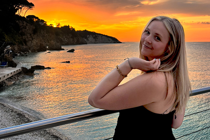 Kaitlyn Klumpp smiles beside a railing overlooking a rocky coastline and the sea at sunset.