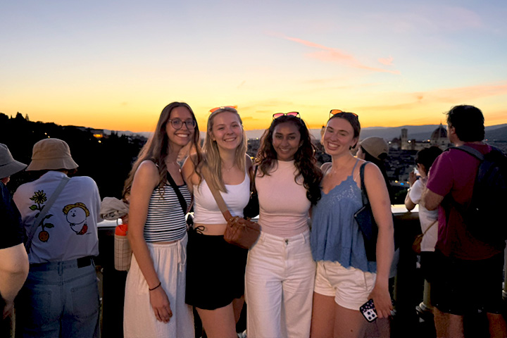 Four students pose together at an overlook as the sun sets over a city skyline.