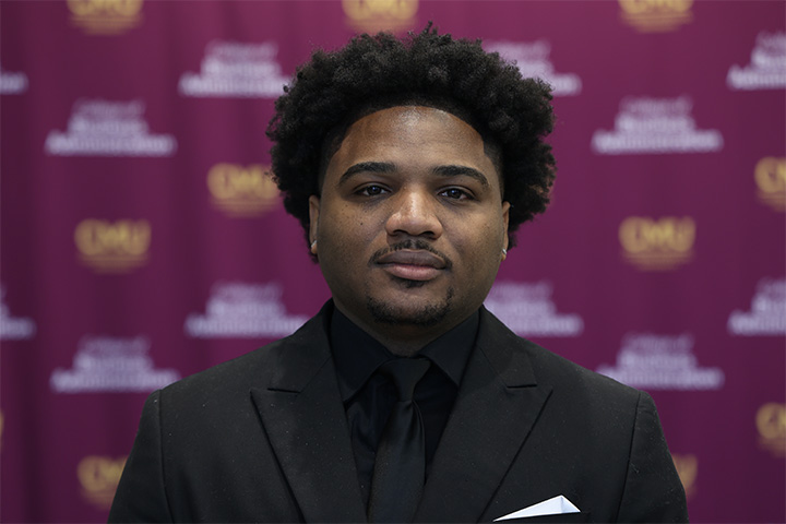 Kalum Gaddis wears a black suit, white pocket square, black shirt and black tie. He has an afro and a goatee. Behind him is a College of Business step and repeat.