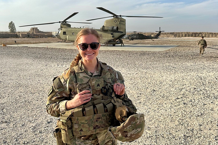 Katie List wears a camouflage uniform and sunglasses and stands in front of an Apache helicopter. The ground is sandy and rocky. Another person is walking away in the background.
