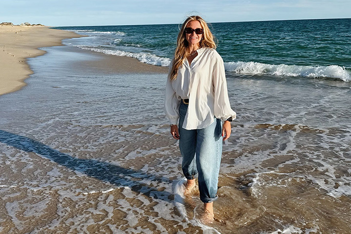 Katie List has long blond hair with beach waves. She's wearing a white button down shirt and jeans that are rolled at the ankles. She's standing in shallow water on a sandy beach and blue waves stretch behind her.