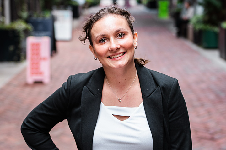 Kayli Kalish stands on a red brick walkway. She has a hand on her hip, head tilted, she smiles at the camera. She's wearing pink pants, a white shirt, and black blazer. Her brown hair is pulled back wish a single curl escaping.