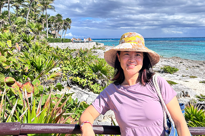 Kun Yang smiling on a coastal boardwalk, wearing a sun hat and lavender dress, with ocean and tropical greenery in the background.