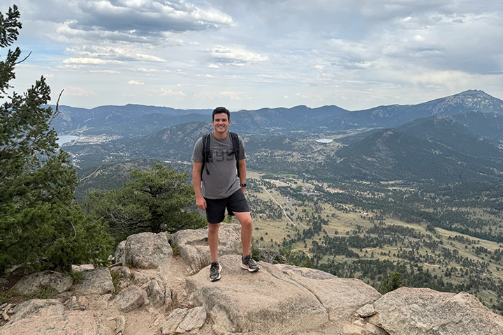 Kurt Paldino stands on a rocky outcropping that overlooks a valley and mountains behind him. He's wearing a black backpack with a gray tshirt and shorts. He smiles at the camera.
