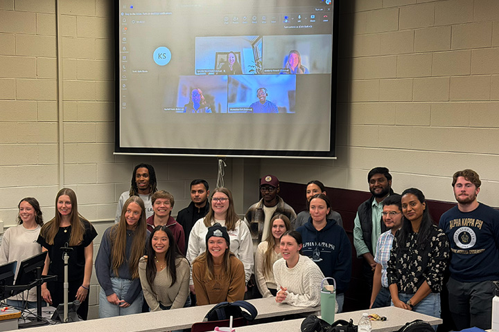 A large group of 18 students from the American Marketing Association and Alpha Kappa Psi pose in front of a pull-down screen with five virtual attendees displayed for their joint La-Z-Boy event.