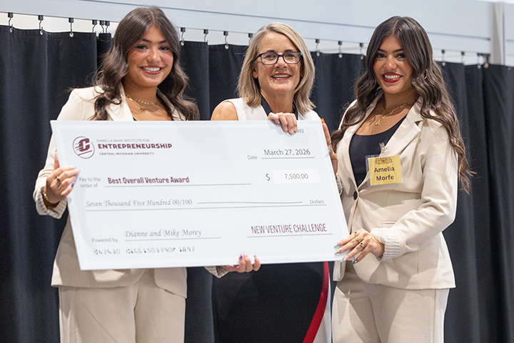 Samantha (left) and Amelia Morfe stand on either side of IBIE Director Julie Messing while holding an oversized ceremonial check for $7,500 after winning first place in Central Michigan University’s New Venture Competition.