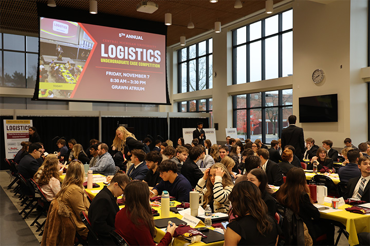 The Grawn Atrium is full of students in business professional clothing. They sit at long tables with gold table cloths. The drop down screen can be seen overhead and it has a slide with the event title and information.
