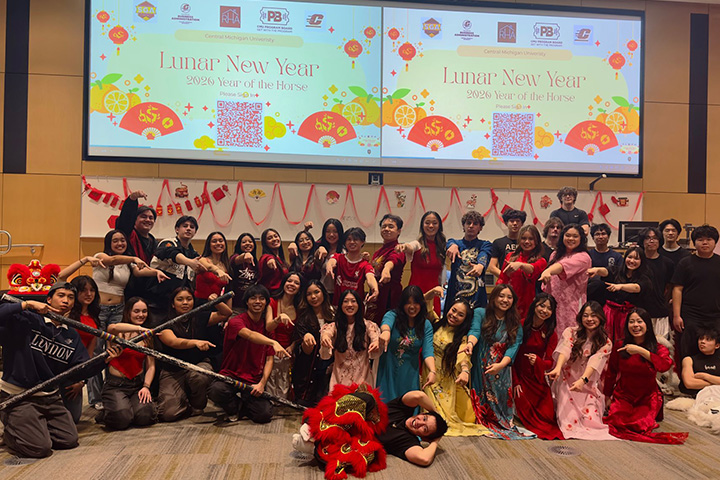 Students dress in traditional Asian dresses, as well as celebration clothes with pops of red, pose in front of two large display screens with Lunar New Year images and the text 