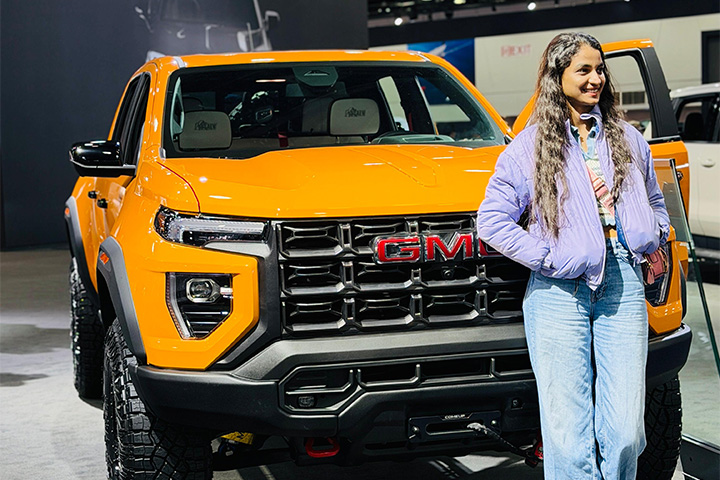 Mounika Gunji stands beside a bright orange GMC pickup truck at an indoor auto show, smiling while wearing a purple jacket and jeans.