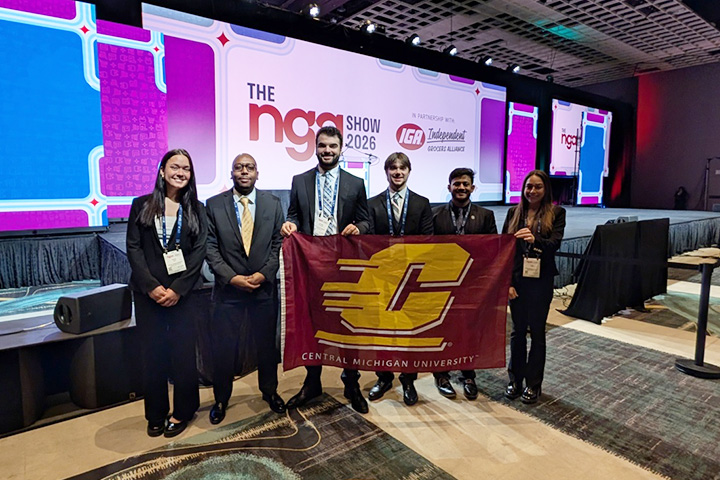 Six CMU logistics students stand with a maroon Action C flag at the National Grocers Association conference where they competed and placed second in a logistics competition.