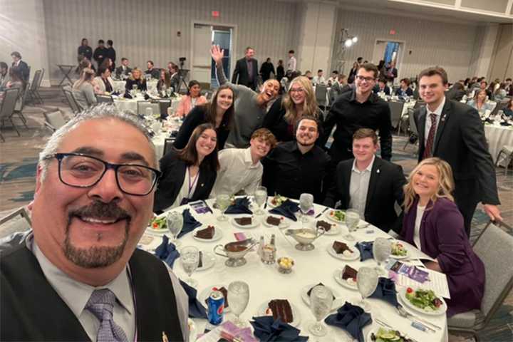Nick Hussein takes a selfie at a PSE event. He has a neat goatee, salt and pepper hair and black glasses. He is wearing his signature vest over a collared shirt and tie. Behind him is a large group of smiling students at a table.
