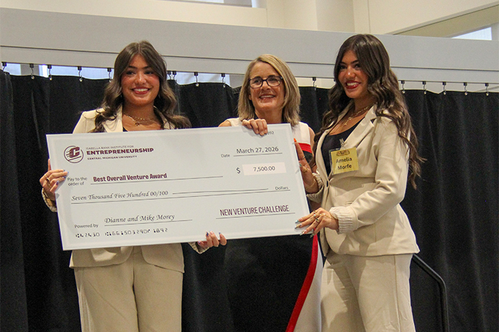 Three individuals stand together holding a large ceremonial check at the New Venture Challenge, including IBIE Director Julie Messing and two student winners.