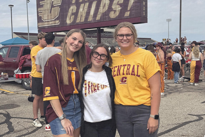 Payton McKay (center) stands with two friends at a CMU football tailgating event. They are dressed in CMU logo gear and all have big smiles. Behind them students stand in the stadium parking lot.
