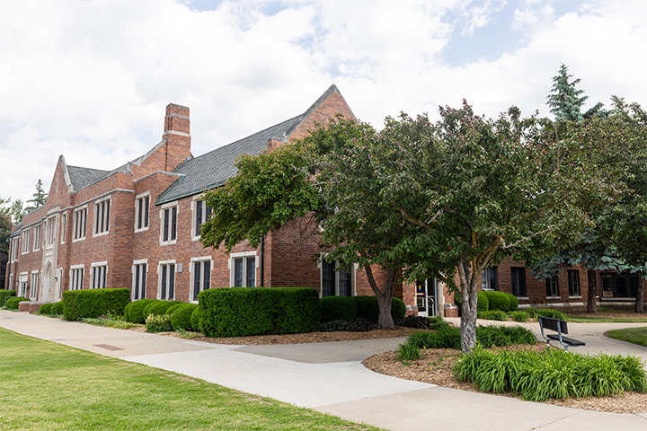 Powers Hall is pictured during the summer. Trees, day lilies and a bench are near the front entrance of the building and green grass is in the foreground.