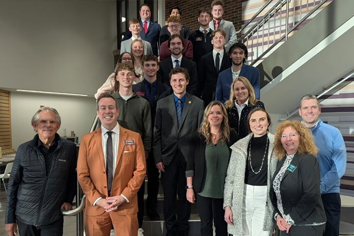 President Neil MacKinnon stands in front of the Grawn atrium staircase with a group of CBA students and supporters. The group is posed together and smiling at the camera. MacKinnon is wearing a burnt orange suit. He's known for his sartorial boldness.