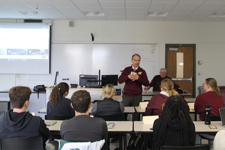 Ricardo Resio wears a maroon CMU pullover while speaking in a class for Dialogue Days. Students at desks in front of him are focused on him as he speaks.