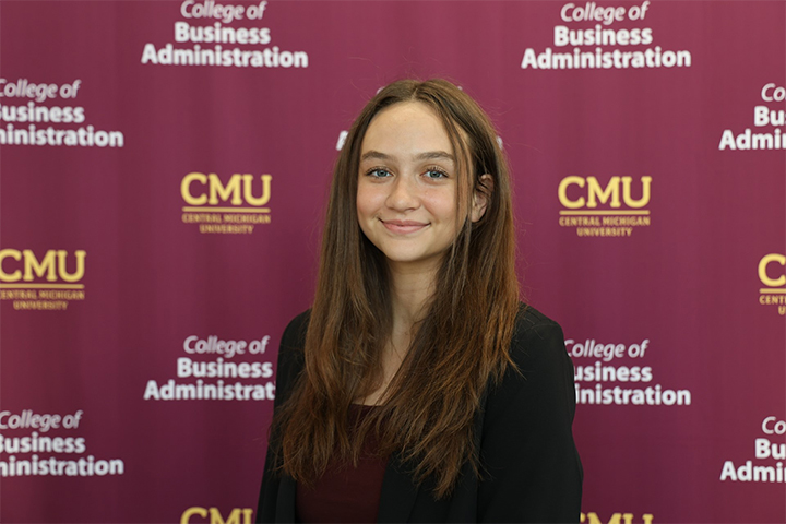 Sarah Firchau stands in front of a maroon College of Business step-and-repeat backdrop. She has long brown hair that is parted in the middle. She wears a black blazer and a deep maroon shirt. She smiles at the camera.