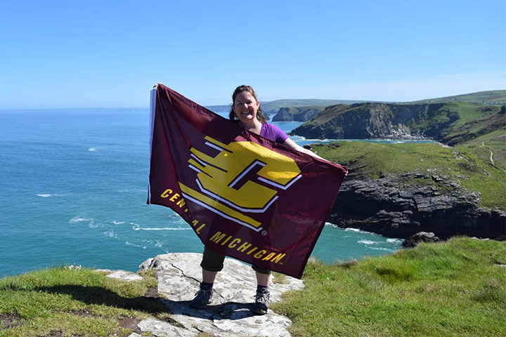 Shelly Bartosek stands on a sunny seaside cliff in the United Kingdom. She holds a maroon CMU flag with a large gold Action C in the center.