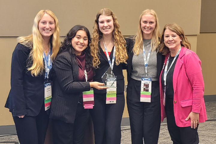 Four female students stand together in a line. Three have long blond hair and one has long dark brown hair. They are all wearing black business suits and lanyards. One holds a crystal trophy. The advisor is at the right side in a bright pink jacket.
